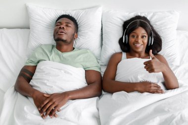 Young african american guy sleeping on bed, cheerful lady in headphones shows thumb up in bedroom, top view