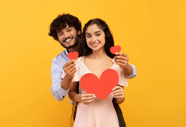 Love is in the air. Romantic indian couple with red paper hearts in hands posing over yellow studio background — Stockfoto