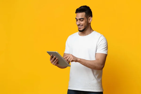 Cheerful arabic guy using digital tablet, yellow background — Zdjęcie stockowe