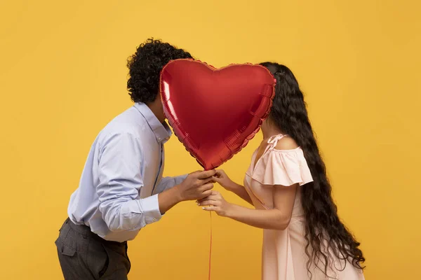 Romantic indian couple kissing behind heart shaped balloon on Valentines Day, posing over yellow background, side view — стокове фото