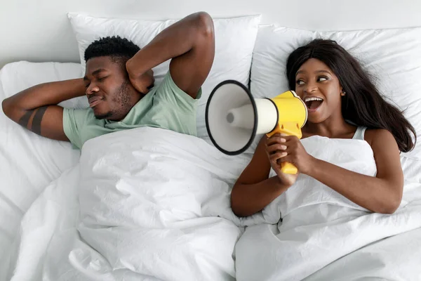 Сумний молодий афроамериканець закриває вуха на білому ліжку, весела жінка кричить на мегафон у спальні. — стокове фото