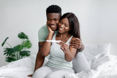 Happy young pretty african american wife and husband on bed in bedroom look at pregnancy test and enjoy good news