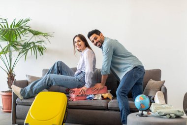 Vacation concept. Excited young spouses trying to close suitcase full of clothes on sofa, preparing for holidays