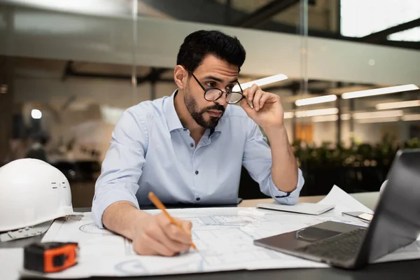 Серйозний зайнятий тисячолітній привабливий арабський інженер в окулярах дивиться на ноутбук і працює з кресленнями проекту — стокове фото