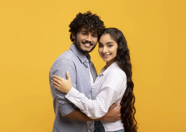 Happy young indian spouses. Portrait of cheerful couple hugging and smiling at camera, standing over yellow background — Φωτογραφία Αρχείου