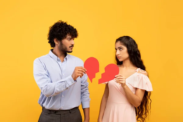Disappointed indian man and woman holding two pieces of broken paper heart standing over yellow background — Φωτογραφία Αρχείου