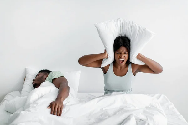 Unhappy young african american lady freaks out, screams, covers ears with pillow on bed, suffers from husband snoring — Fotografia de Stock