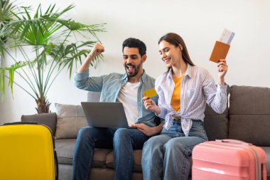 Excited tourists couple booking hotel room online, using laptop and credit card, sitting on sofa with suitcases