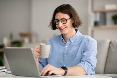 People and technology concept. Young guy using laptop and holding cup, working and drinking hot coffee