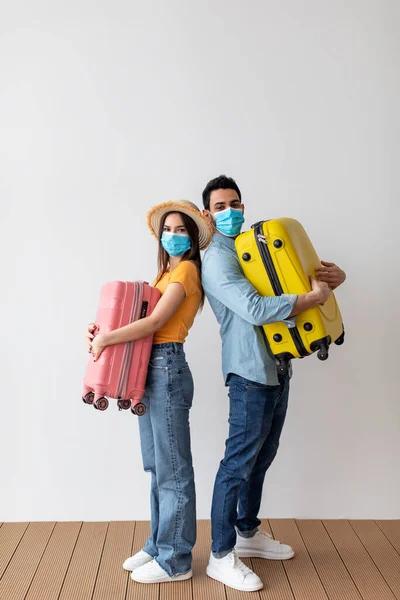 Tourists couple in face masks embracing their suitcases, standing over light wall. Vaccinated spouses ready for vacation — 스톡 사진