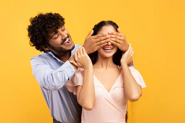 Conceito de surpresa romântico. Retrato de cara indiano feliz cobrindo seus olhos bonita namorada de volta, fundo amarelo — Fotografia de Stock