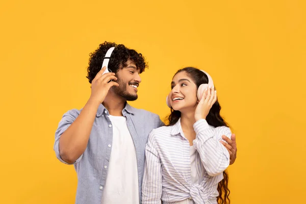 Loving indian couple in headphones listening to music, hugging and looking at each other over yellow studio background — ストック写真