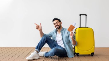 Happy arab man holding plane tickets and passport, pointing at free space, sitting near suitcase over light wall
