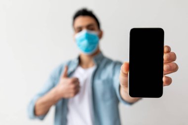 Arap adam akıllı telefonlu boş ekran gösteriyor ve baş parmağını kaldırıyor, koruyucu yüz maskesi takıyor, seçici odaklanıyor.
