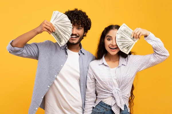 Amantes indios millennials ricos escondiendo caras detrás del efectivo, sosteniendo racimos de dólares por caras sobre fondo amarillo —  Fotos de Stock
