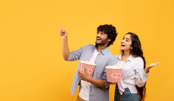Happy indian couple eating popcorn, cheerful guy pointing at free space for advert or text, panorama — 图库照片