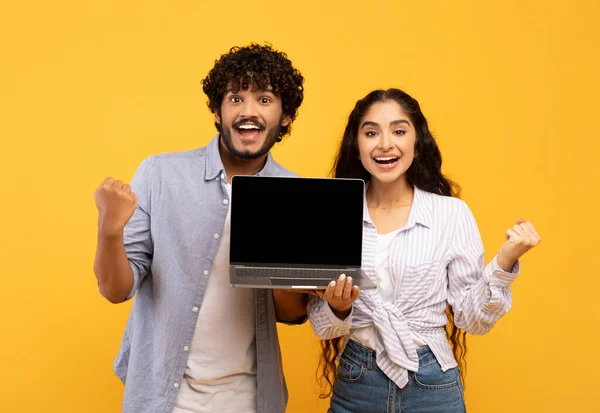 Yes. Portrait of happy indian couple holding laptop with blank screen, shaking clenched fists, mockup, yellow background — Foto Stock