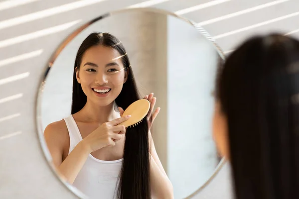 鏡の前に立つ陽気なアジアの女性のブラッシング髪 — ストック写真
