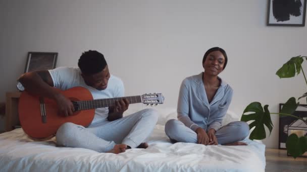 Hiburan keluarga. Happy african american guy playing guitar, his happy girlfriend singing, sitting together on bed — Stok Video