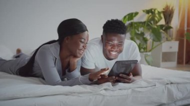 Mutlu anılar. Genç Afro-Amerikan çifti yatakta dinleniyor ve sabahları dijital tablette fotoğraf izliyorlar.