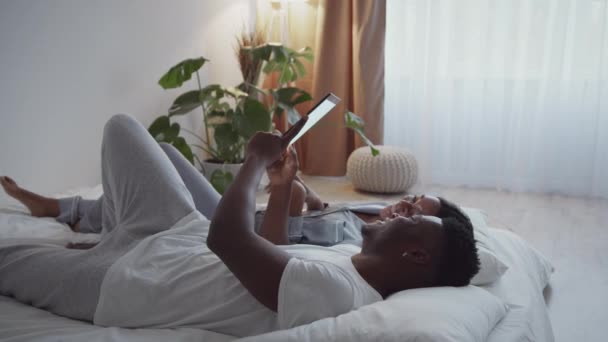 Giovane amorevole coppia afroamericana in pigiama sdraiato a letto a guardare la tv online su tablet digitale, a riposo al mattino — Video Stock