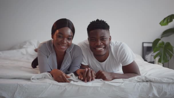 Jonge liefdevolle Afrikaanse Amerikaanse echtgenoten die elkaars hand vasthouden, op bed liggen en thuis naar de camera glimlachen, shot volgen — Stockvideo