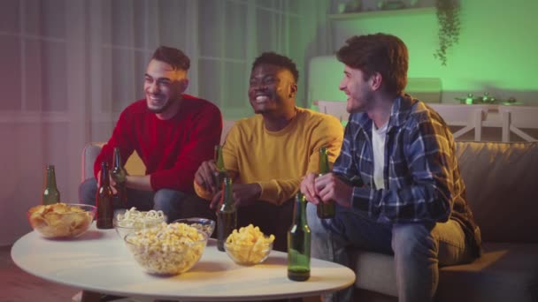 Tři multietničtí kluci si spolu užívají páteční večer, dívají se na televizi a jásají s pivem doma v neonových světlech — Stock video