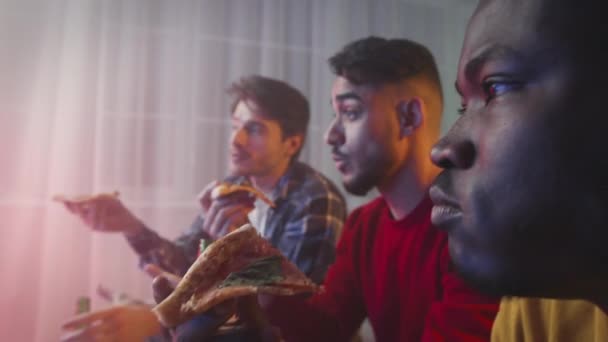 Male singles party. Zoom in portrait of happy diverse men eating fresh pizza and watching tv at home in neon lights — Video Stock