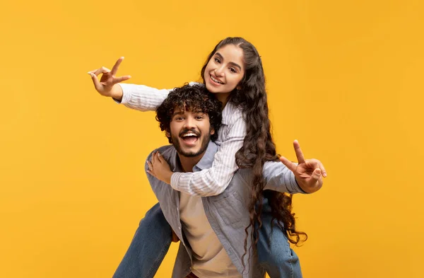 Romantik Hintli çift birlikte eğleniyor, V işaretini gösteriyor, erkek kız arkadaşını sırtında taşıyor, sarı arka plan. — Stok fotoğraf