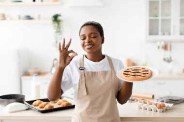 Satisfied millennial pretty black lady in apron enjoys aroma of fresh tasty pie in modern kitchen interior, free space