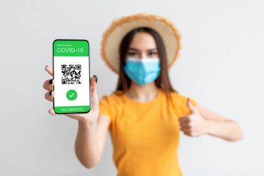 Elektronik sağlık pasaportu. Yüz maskesi takan ve elinde dijital e-pasaport olan akıllı telefon tutan genç bir kadın.