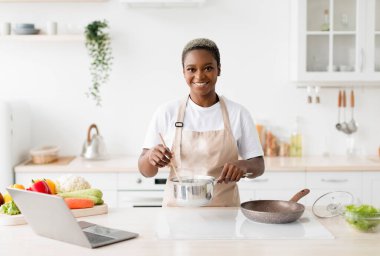 Happy young cute black lady in apron prepares lunch on table with pc in minimalist kitchen interior, free space