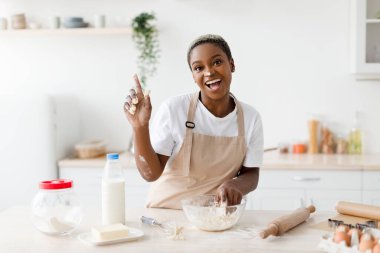 Laughing millennial african american woman in apron prepares dough for baking and raises finger up