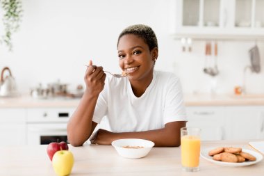 Smiling young attractive black woman eating porridge sits at table with cookies, juice and apples