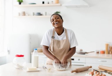 Happy glad laughing young attractive black lady in apron prepares dough for baking in modern kitchen interior