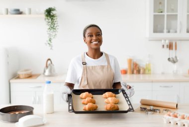 Satisfied young cute black woman in apron shows ready croissants in modern kitchen interior