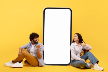 Happy indian couple sitting near big smartphone with blank white screen, demonstrating copy space for app or ad