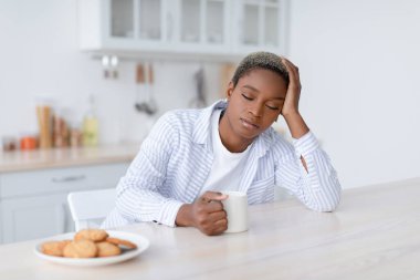 Tired sleepy young attractive black woman with cup of drink sits at table with cookies in scandinavian kitchen interior