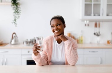 Smiling glad young attractive black lady holds glass of clean water, pensive and look at free space