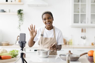 Happy young pretty black woman blogger in apron prepares dinner, shows dish, waves hand, shoots video for blog