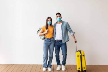 Coronavirus and tourism. Young couple in medical masks ready for vacation during pandemic, standing with baggage