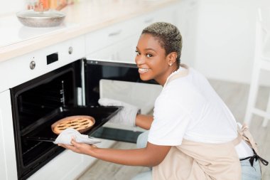 Cheerful young pretty black lady with short haircut takes out pie from oven in modern minimalist kitchen interior