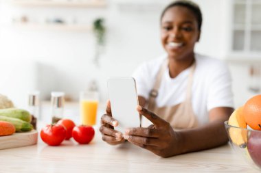 Smiling young pretty black lady in apron reads message on phone chatting in social networks at table with vegetables