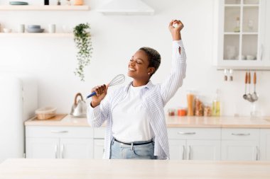 Cheerful millennial pretty black female singing at appliance as imaginary microphone, dancing