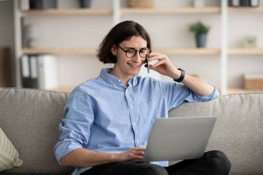 Home office concept. Excited guy talking on smartphone and using computer laptop, working remotely from home