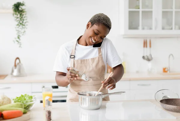 Cheerful millennial pretty black female in apron preparing lunch talk by phone in scandinavian kitchen interior — Stock Photo, Image