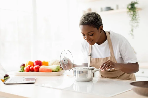 Cheerful young pretty black female in apron prepare lunch and smell dish in minimalist kitchen interior — Stock Photo, Image