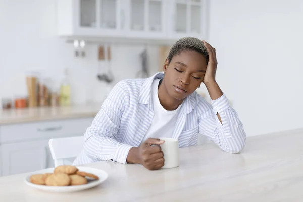 Tired sleepy young attractive black woman with cup of drink sits at table with cookies in scandinavian kitchen interior — Zdjęcie stockowe