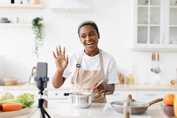 Happy young pretty black woman blogger in apron prepares dinner, shows dish, waves hand, shoots video for blog — Stock Photo, Image