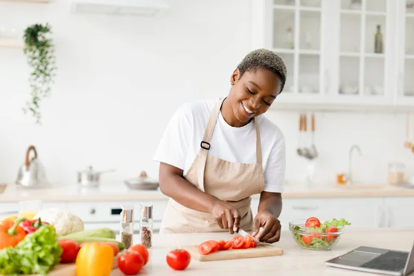 Smiling millennial pretty black female in apron prepare healthy lunch from organic vegetables, cuts tomatoes — Stockfoto